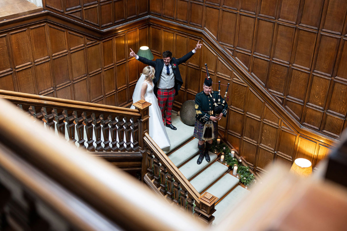 Groom raises his arms as he and the bride walk downstairs behind a piper