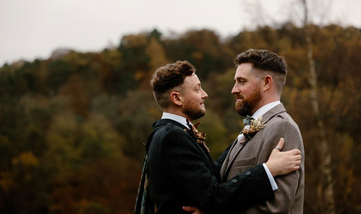 two grooms wearing tweed jackets standing face to face with autumn landscape in background at Neidpath Castle