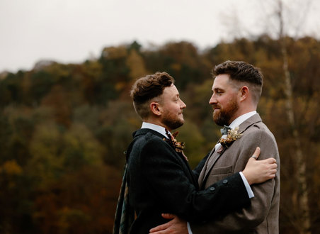 two grooms wearing tweed jackets standing face to face with autumn landscape in background at Neidpath Castle
