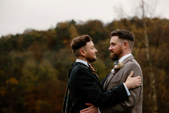 two grooms wearing tweed jackets standing face to face with autumn landscape in background at Neidpath Castle