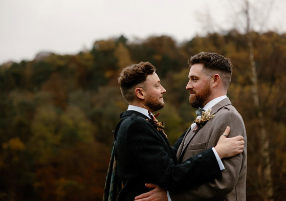two grooms wearing tweed jackets standing face to face with autumn landscape in background at Neidpath Castle