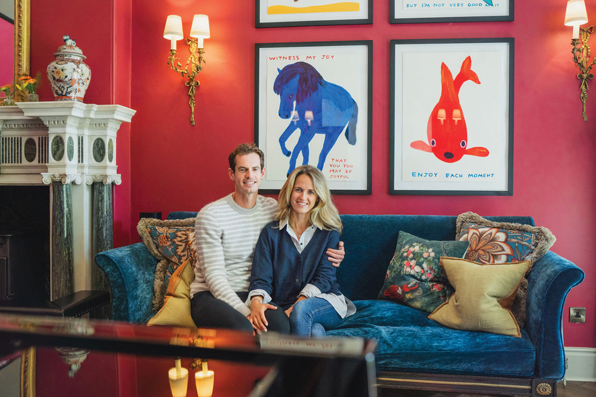 Couple sitting together on a teal velvet sofa in a red-walled living room with framed artwork and ornate lighting behind them
