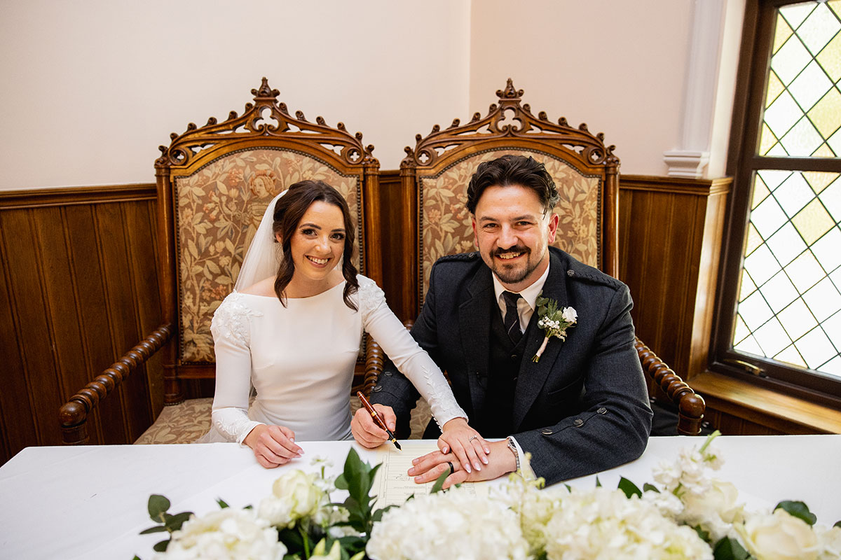 Bride and groom sit together signing the marriage certificate, smiling for the camera