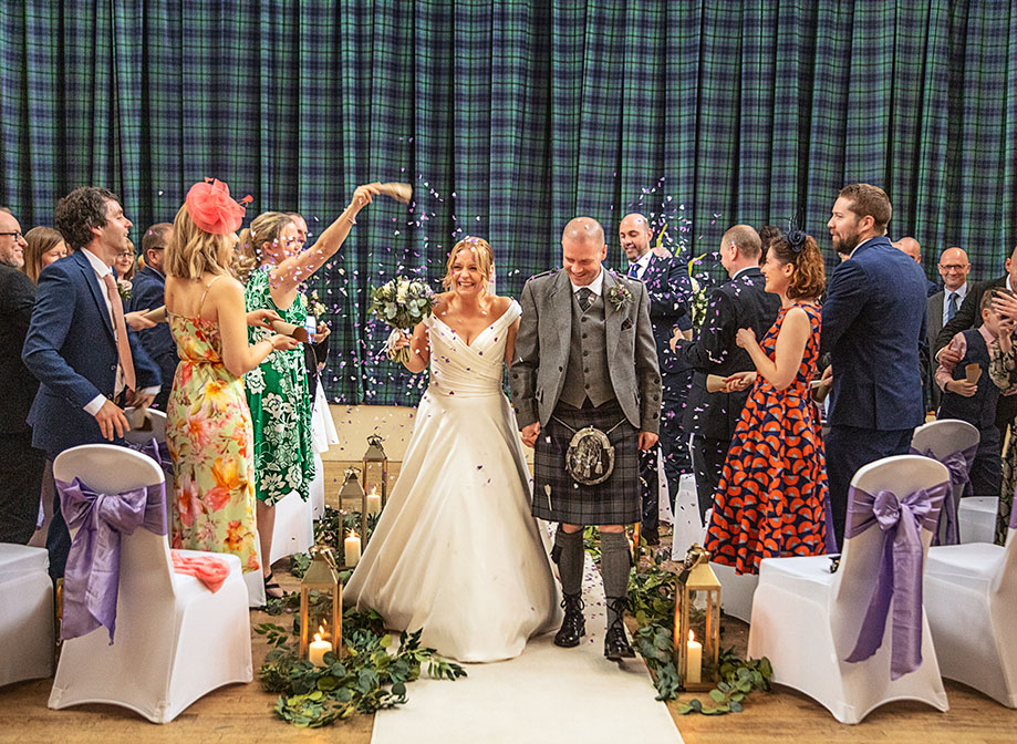 Wedding guests throw purple confetti over a bride and groom as they walk down a white aisle