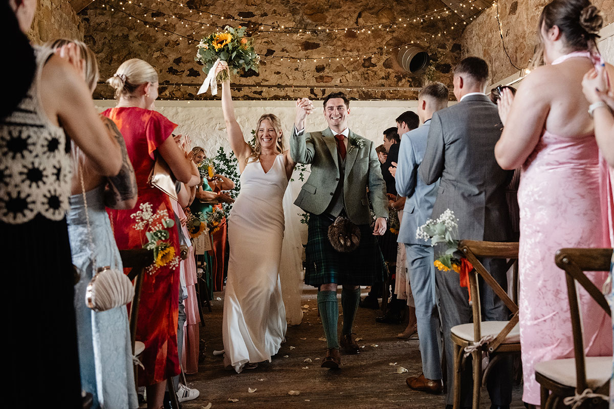 Bride and groom walking back up the aisle, holding hands and celebrating as guests cheer inside the ceremony barn