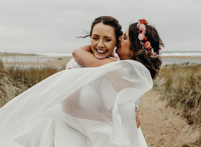two brides on the beach wearing white dresses, one wearing a pink flower crown