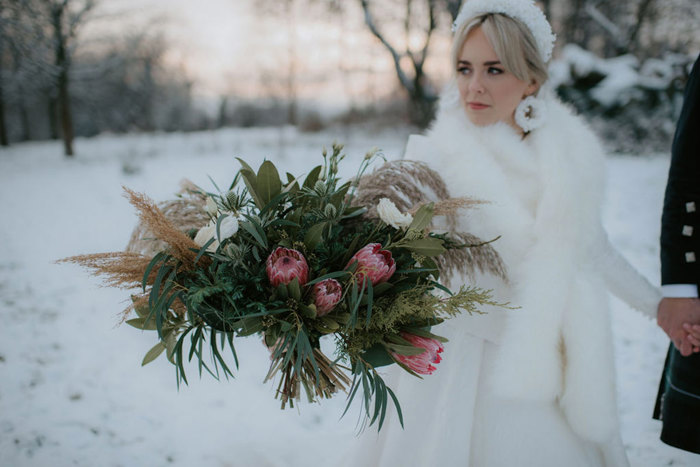 A bride in the snow wearing a white dress and white fur wrap holding a bouquet with pink flowers