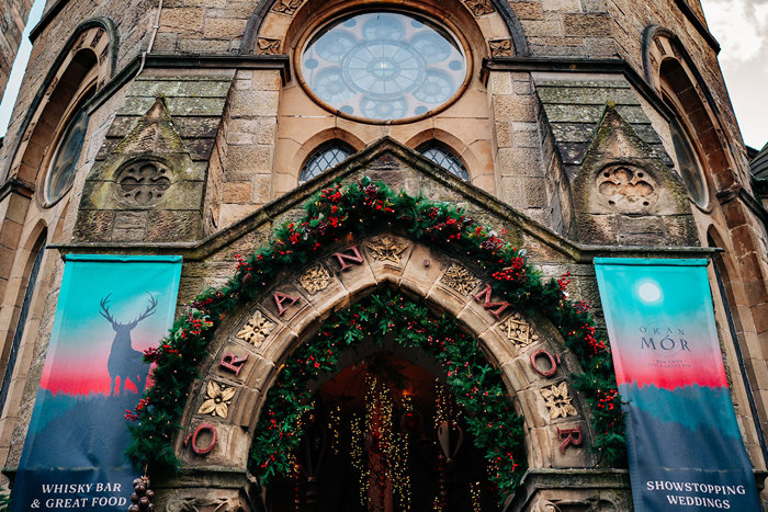 the arch entrance of a tall church-like building labeled Oran Mor covered in Christmas greenery, red berries and twinkling lights with two canvas banners also on either side, advertising the whisky bar, great food and showstopping weddings