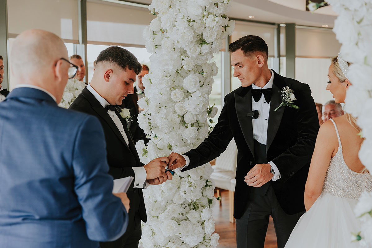 groom in black tux stands at white flower arch, retrieving a ring from another man in tux, as blonde bride stands across from him