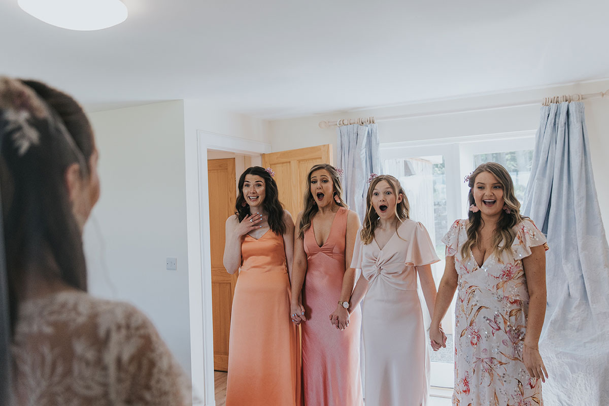 bridesmaids wearing different coloured pastel dresses look surprised as they do a first look with a bride