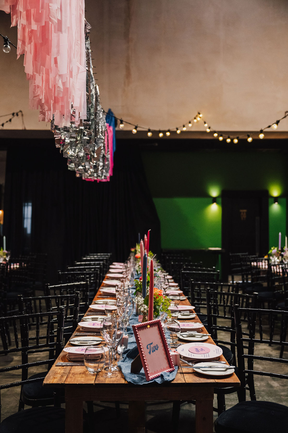 Long wooden dining table set for a wedding meal, styled with colourful taper candles, bright florals and pink stationery.