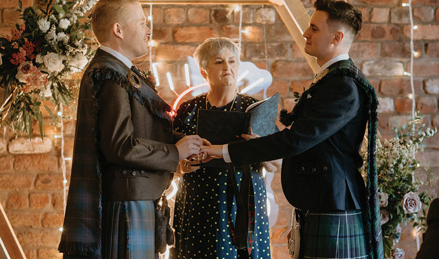 Two men in kilts exchanging rings during a wedding ceremony in front of a wooden hexagon with neon sign, with officiant in background