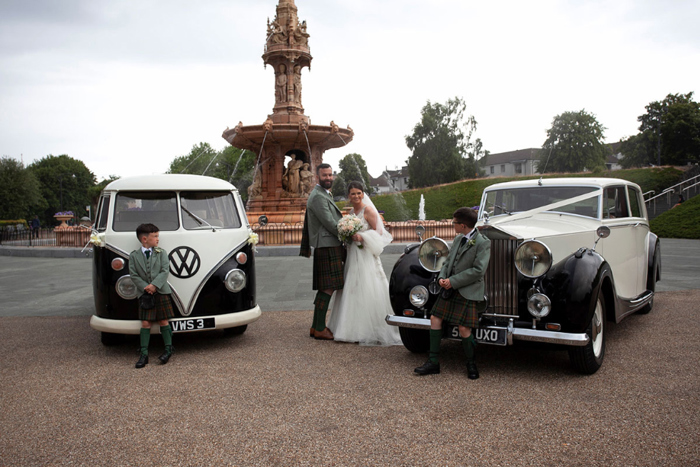 A bride and groom stand in between a Volkswagen camper van and a vintage car 