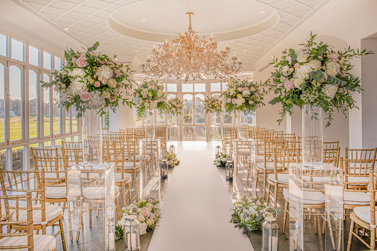 Elegant wedding ceremony setup with floral arrangements, gold chairs and chandelier in a bright luxury venue