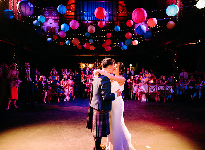 a bride and groom kiss on a dance floor under colourful lanterns as guests seated at tables look on