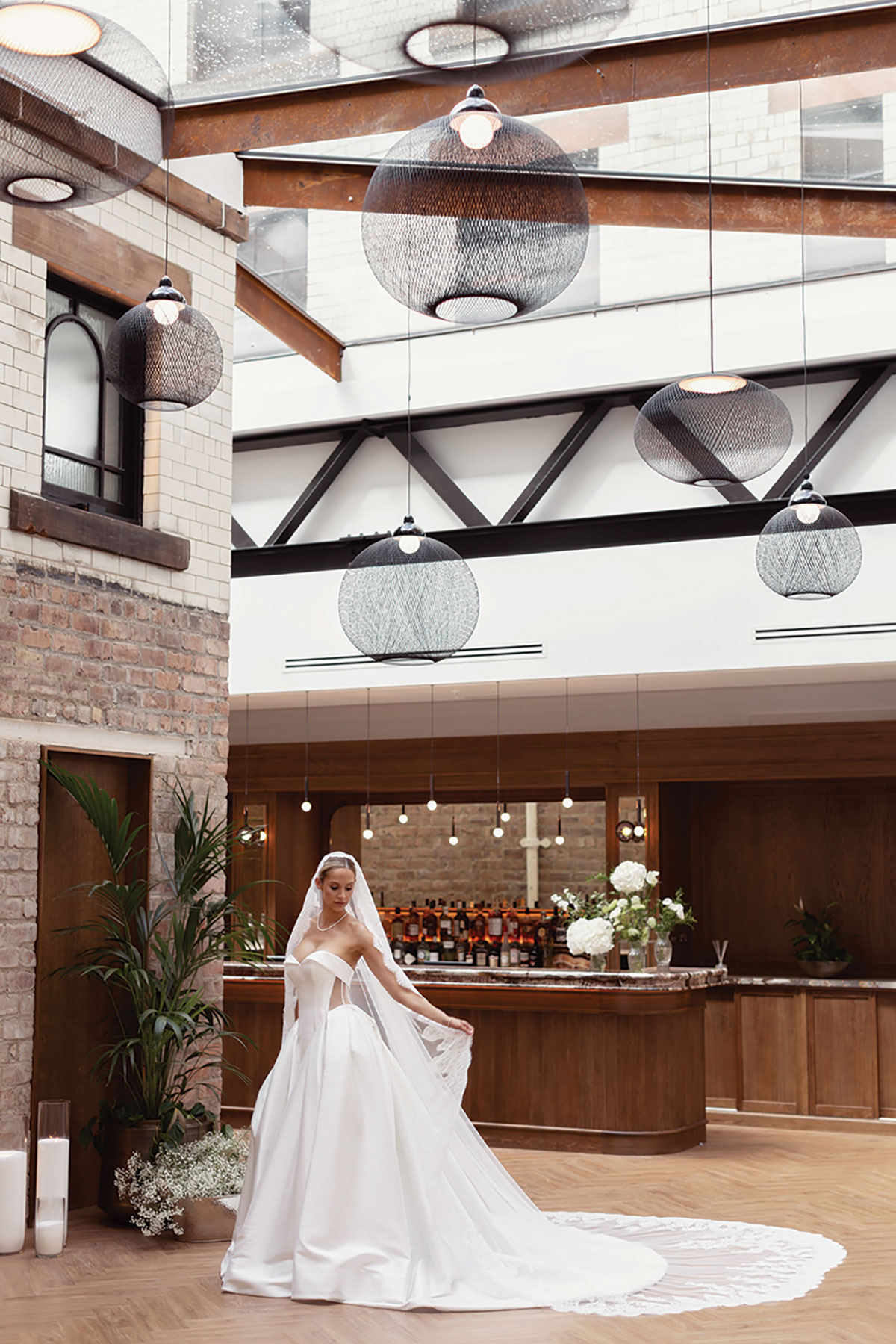 Bride in strapless ballgown wedding dress with lace veil poses in industrial-style venue bar area with exposed brick and glass ceiling