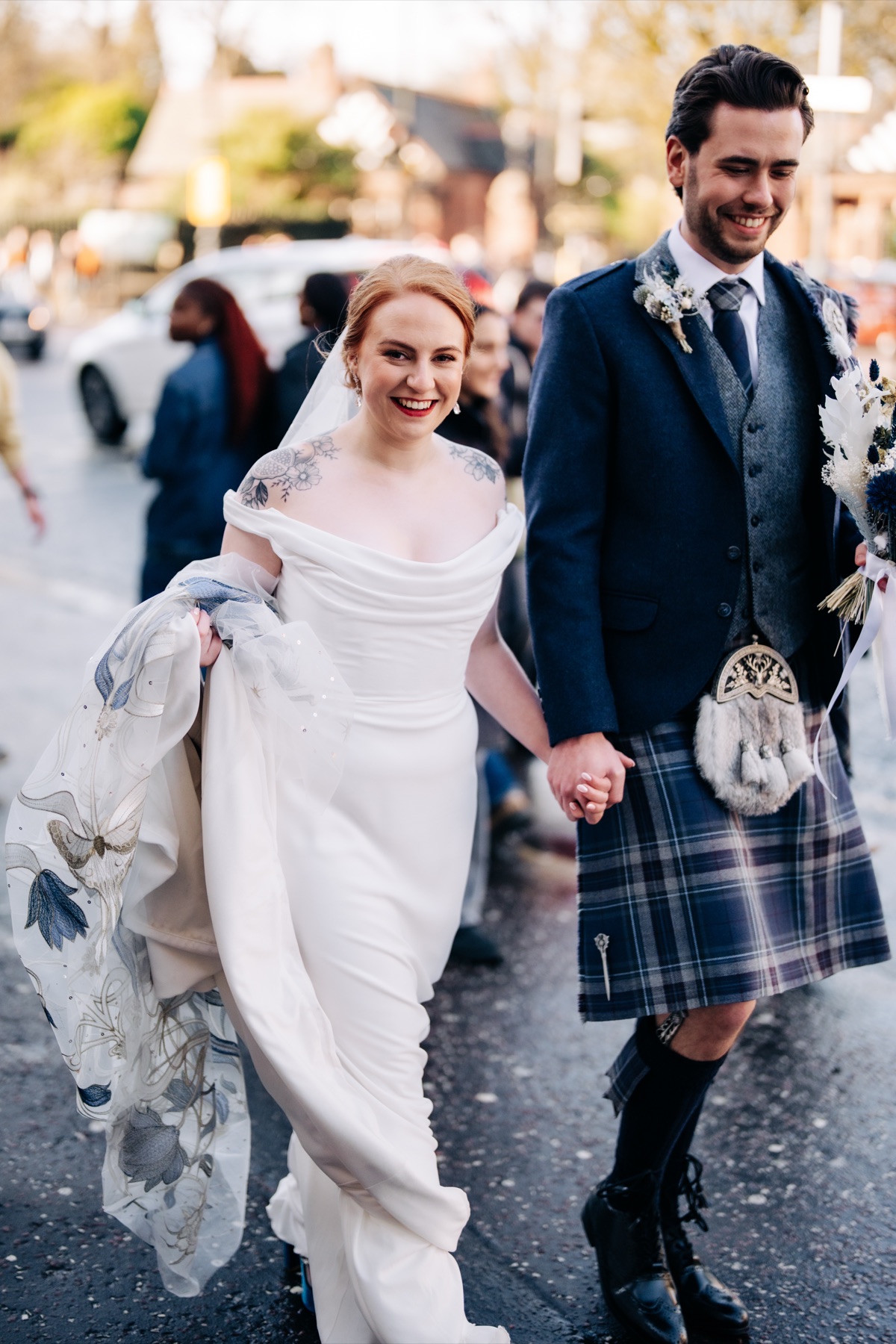 Robyn and James walk hand in hand along Great Western Road following their wedding at Òran Mór in Glasgow, wearing traditional Scottish wedding attire.