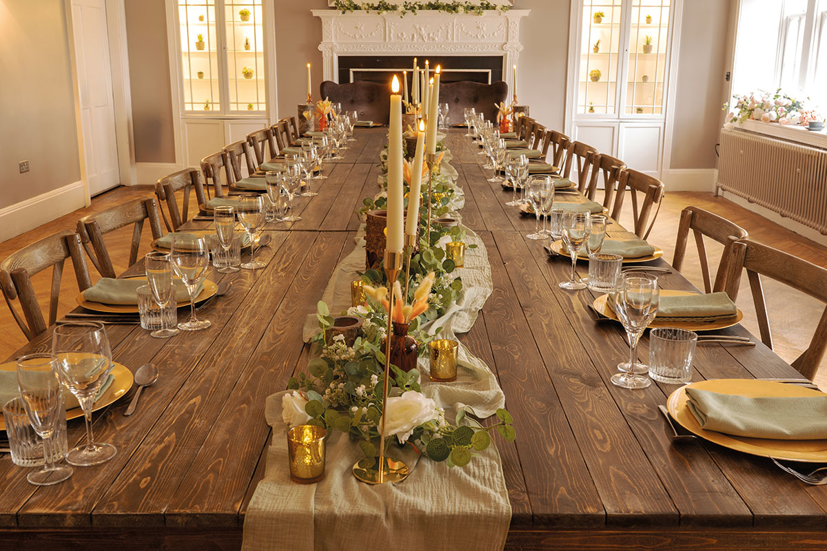 long wooden banquet table with pale green runner down the centre and covered in candlesticks, greenery and surrounded on both sides by place settings