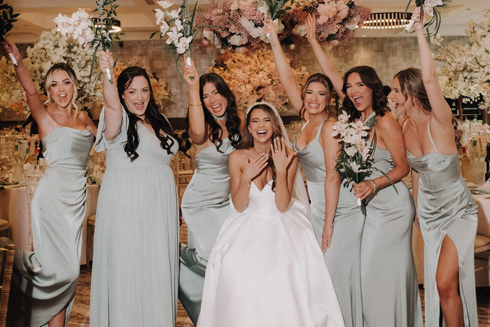 a bride showing off a wedding ring with a row of bridesmaids wearing pale green dresses holding their bouquets in the air.