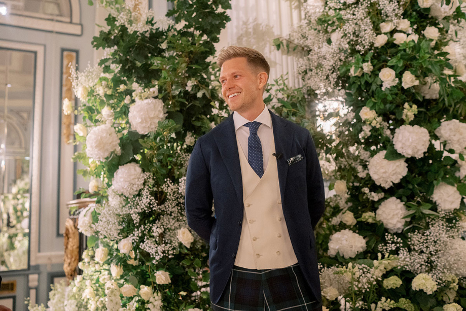 Groom waits for bride at the top of the aisle with white flower installation behind him