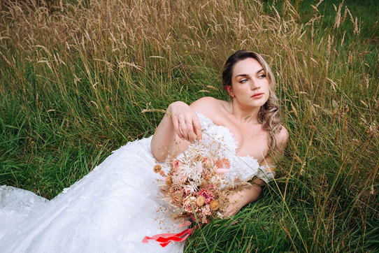 bride in off the shoulder dress lies in a field holding a pink and neutral coloured dried bouquet