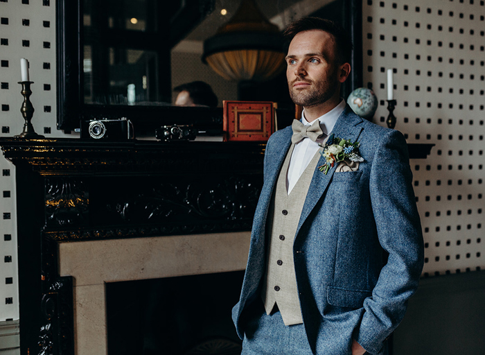 a groom wearing a blue tweed suit standing by a dark wood ornate fireplace with mirror overhanging and black and white checked wall in background