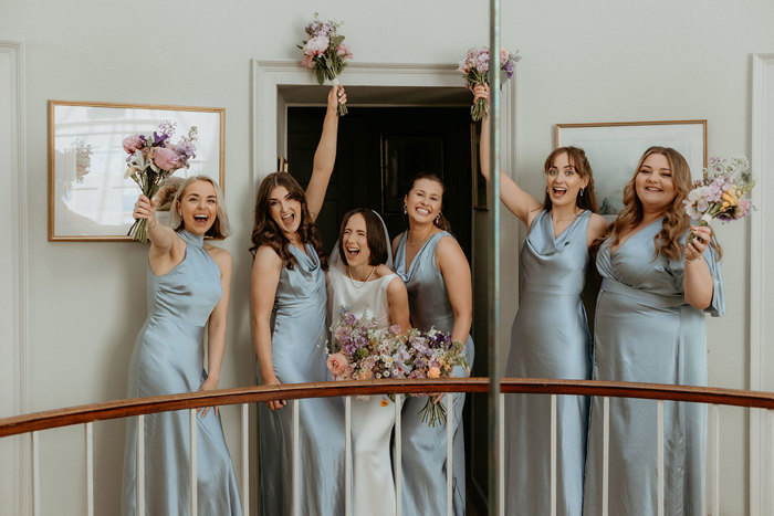 five bridesmaids wearing blue Six Stories dresses and bride pose for a photo behind a white and wooden rail during a wedding at the Byre at Inchyra