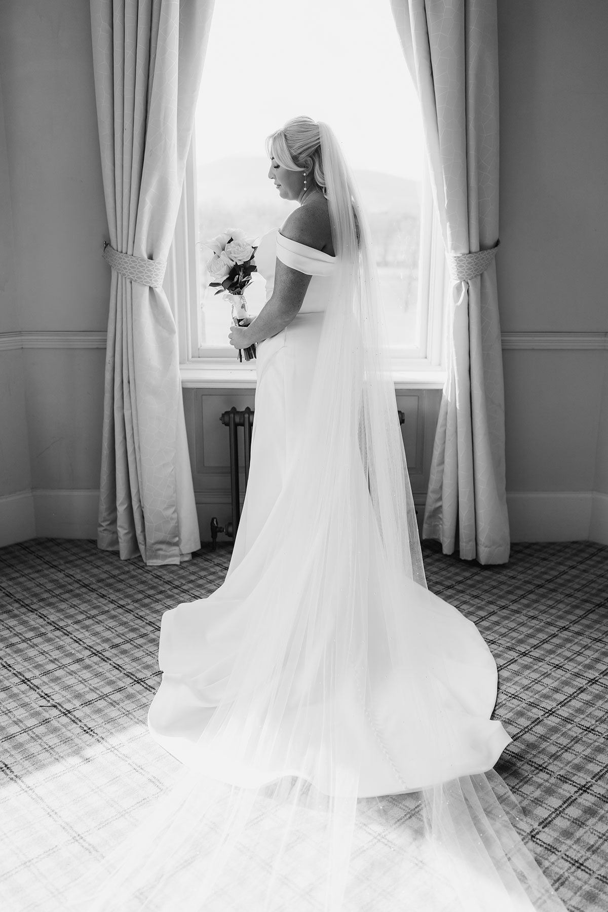 Black and white bridal portrait of the bride holding her bouquet by the window, showcasing her long veil and off-the-shoulder gown
