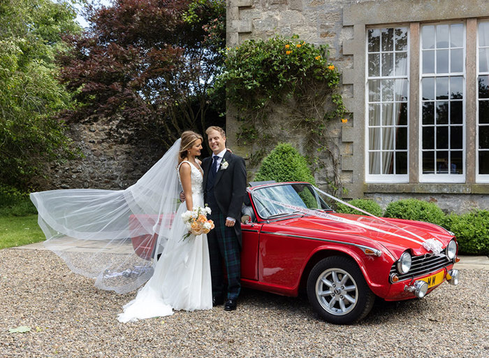 Bride And Groom Standing Next To Red Convertible