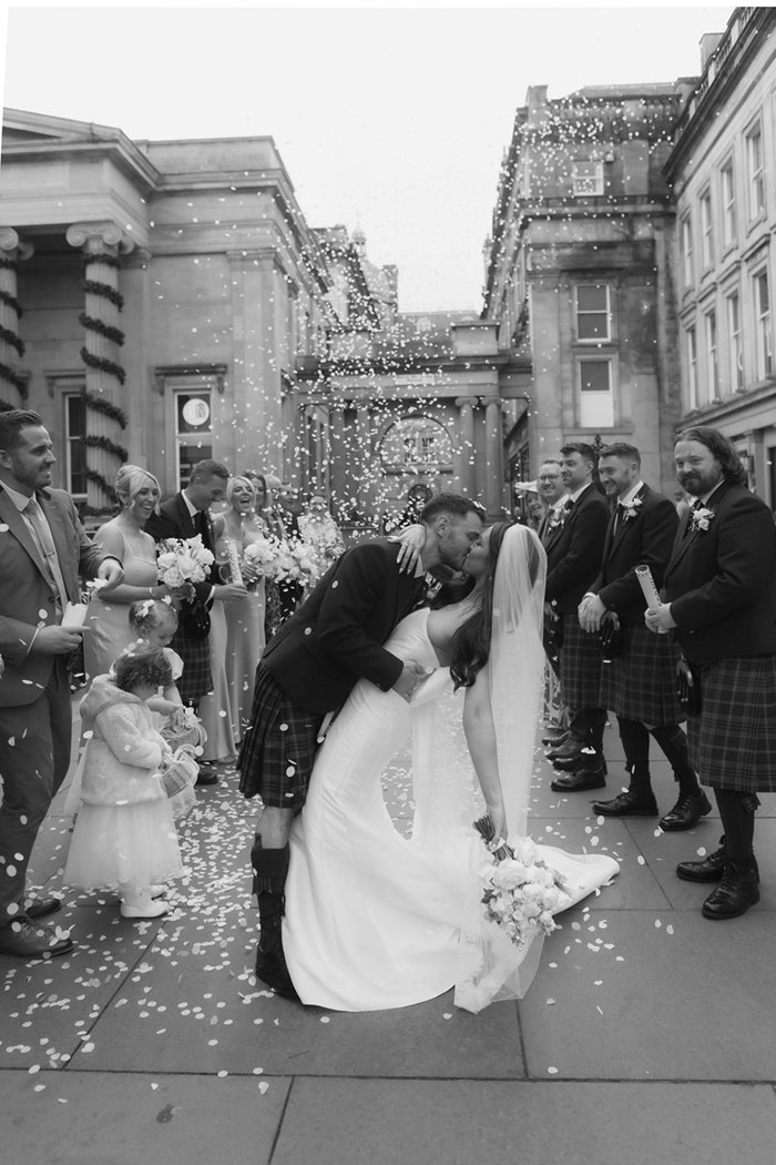 A black and white photo of a bride and groom kissing as confetti is thrown over them by their guests