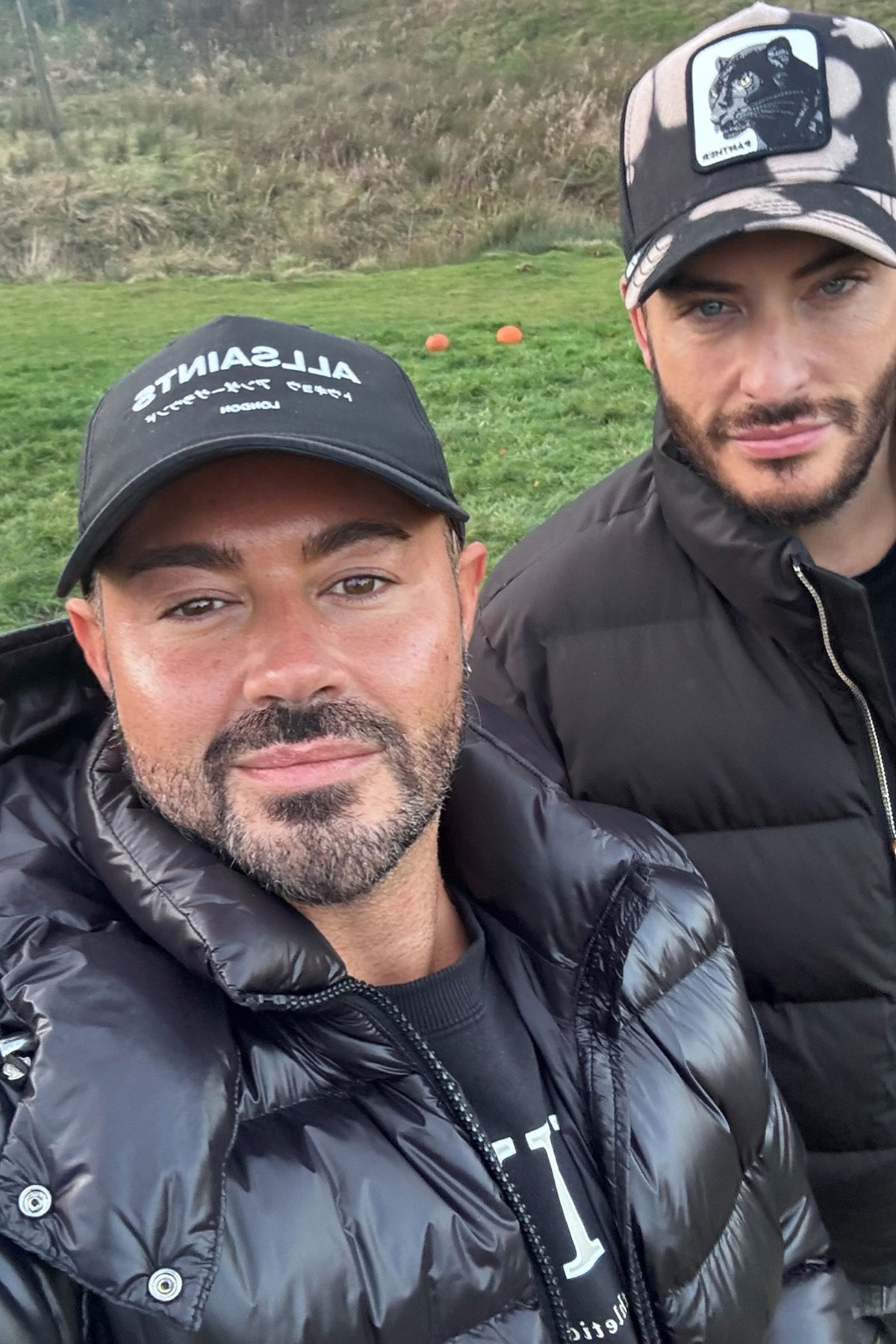 Two men outdoors taking a close-up selfie, both wearing black puffer jackets and baseball caps, standing on grassy terrain