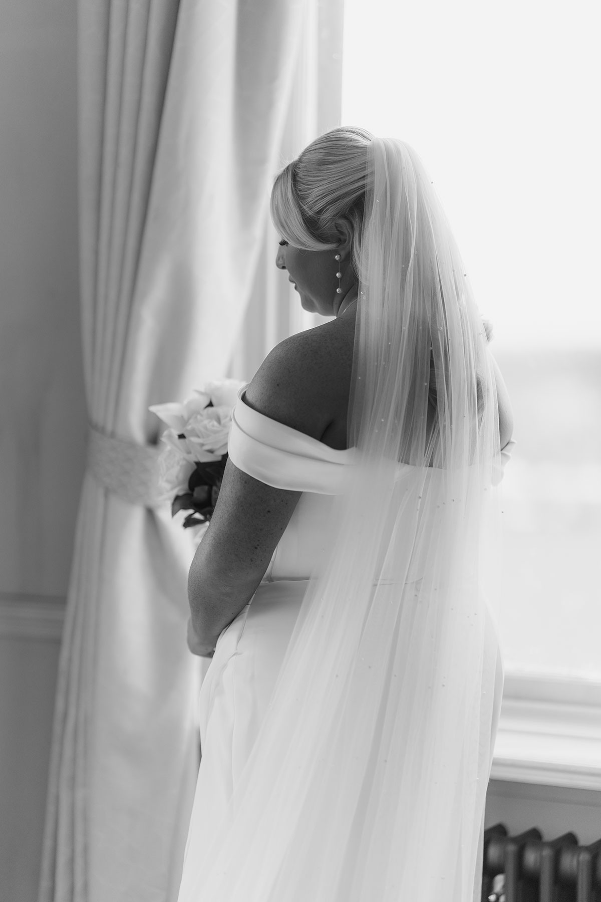 Black and white bridal portrait from behind, showcasing the bride's pearl-detailed veil and elegant off-the-shoulder gown