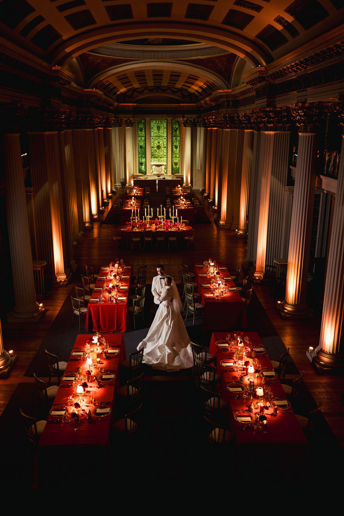 Bride and groom standing among candlelit reception tables dressed in deep red linens inside a grand hall with tall columns and dramatic lighting