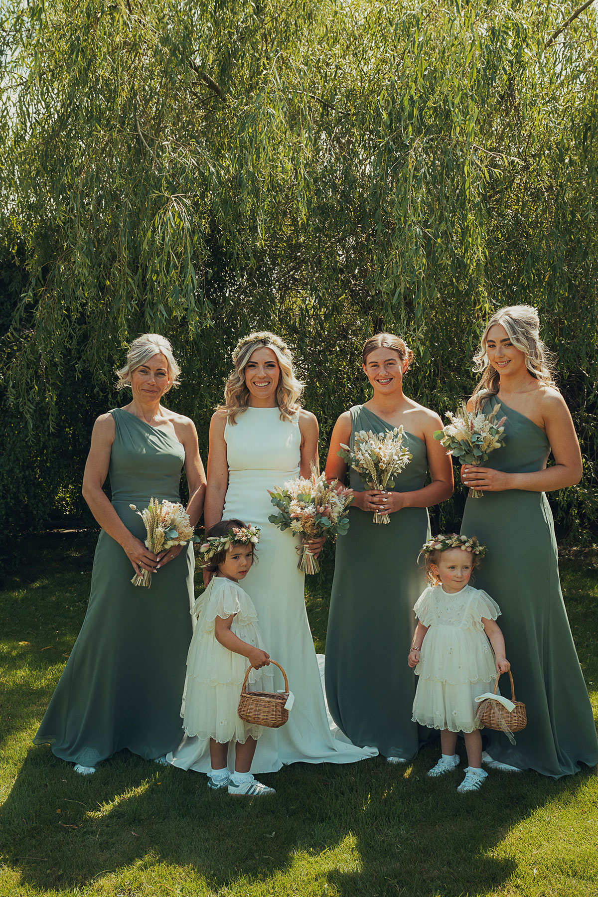Bride stands smiling with three bridesmaids in sage green dresses and two flower girls in white holding baskets, all posing outdoors beneath a tree