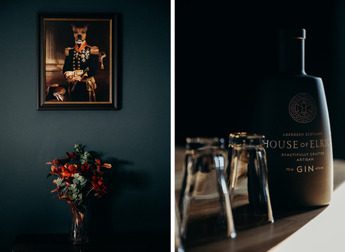 a bunch of red and orange flowers in a vase against a dark teal wall and below a quirky painting of a dog wearing a military outfit on left. A close up of a bottle of House of Elrick gin and shot glasses on right