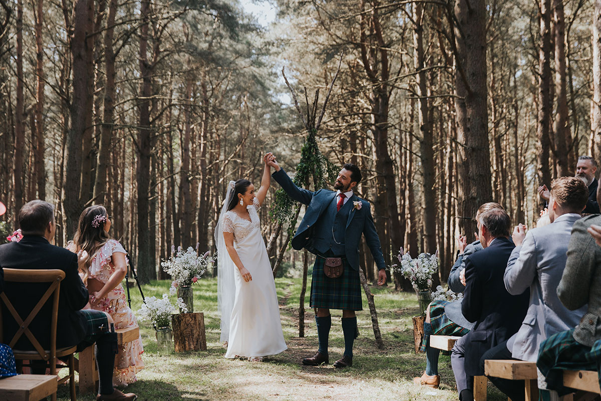bride and groom hold hands in the air in front of wedding guests in a woodland ceremony
