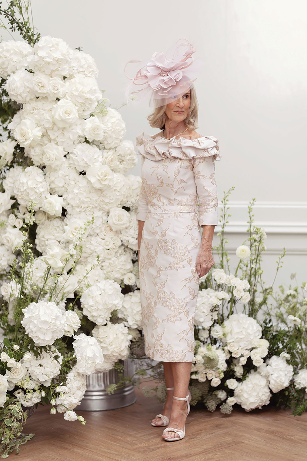 Mother of the bride in floral dress and pink fascinator stands by white rose and hydrangea flower installation