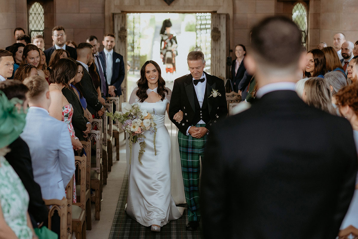 Bride walking down the chapel aisle at Rosebery House and Steading, Midlothian, escorted by her father towards the groom during Scottish wedding ceremony.