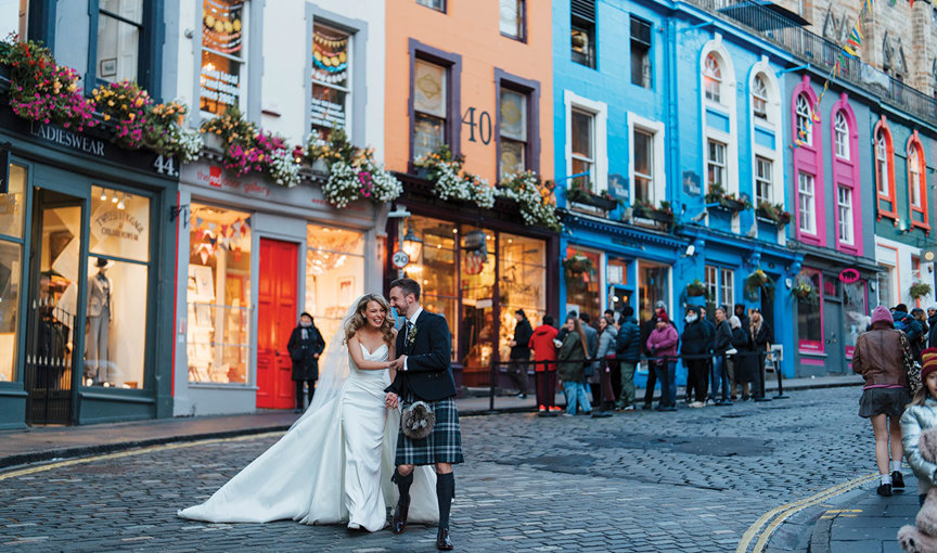 a bride and groom walking on a cobbled street with tall colourful buildings behind them. Victoria Street in Edinburgh