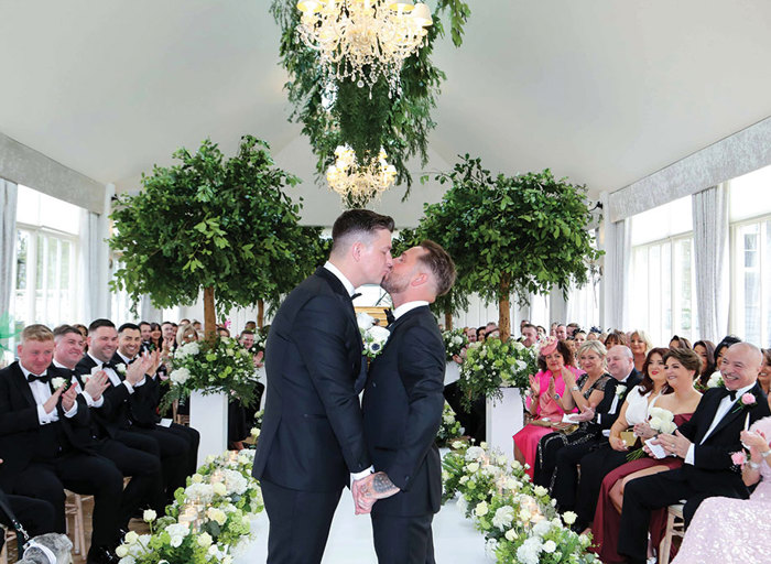 two grooms hold hands and kiss as rows of seated wedding guests either side applaud and smile. There are trees either side of the white aisle and white and green floral arrangements on the floor. There's a large green floral arrangement and two chandeliers hanging from the ceiling