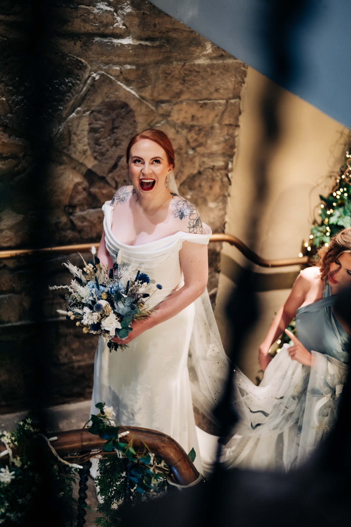 Bride Robyn reacts excitedly before her wedding ceremony at Òran Mór, holding her blue-and-white bouquet on a stone staircase.