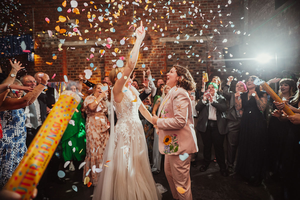 Brides celebrating on the dancefloor under falling confetti as guests cheer and confetti cannons go off around them.