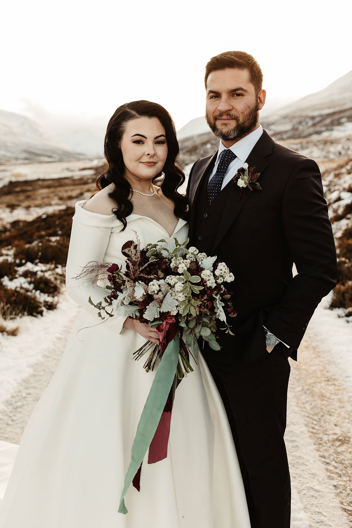 Bride and groom standing on a snow-covered path in the Highlands, holding a winter bouquet against the hillside backdrop
