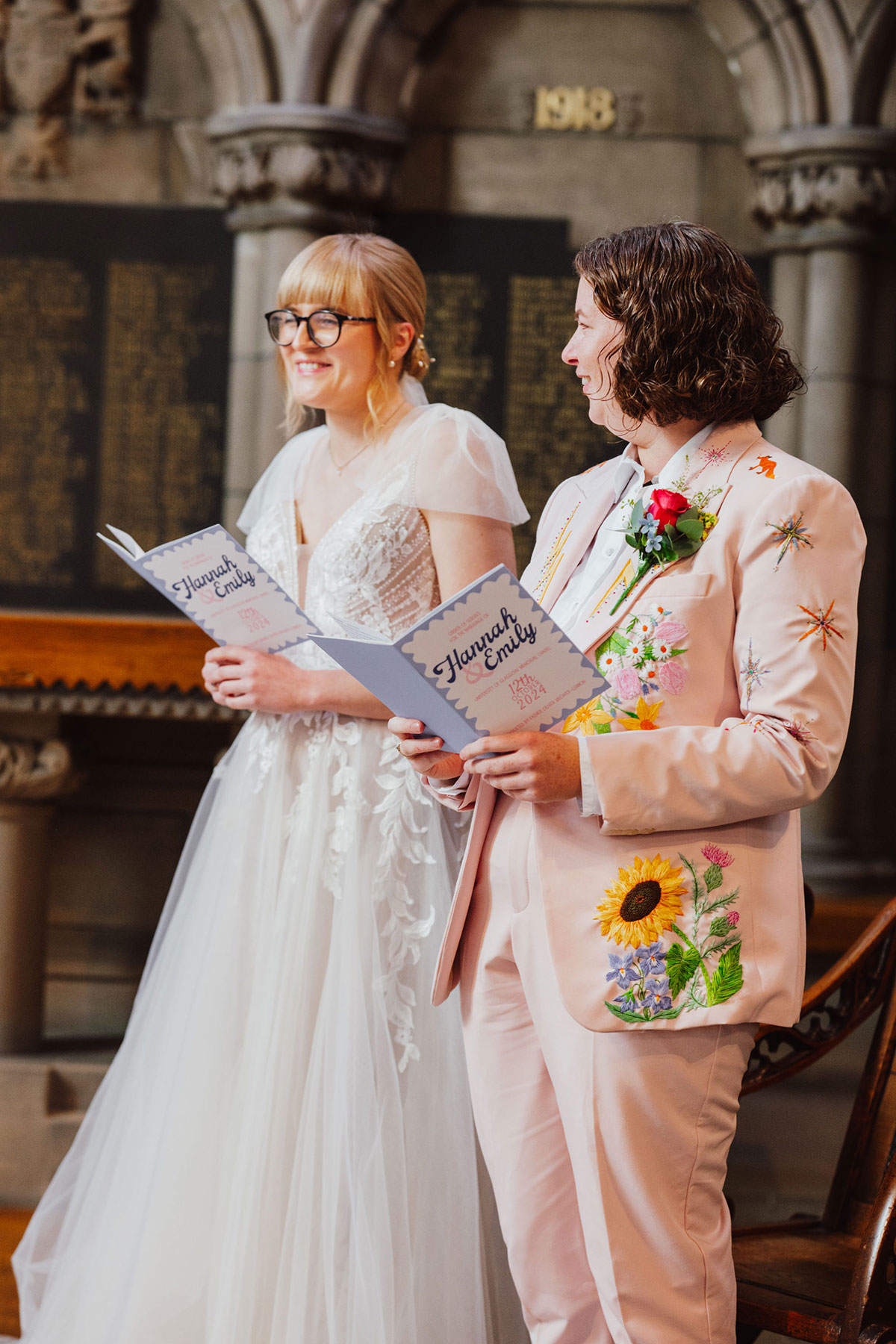Two brides standing side by side during their ceremony, holding matching illustrated orders of service featuring their names and wedding date.
