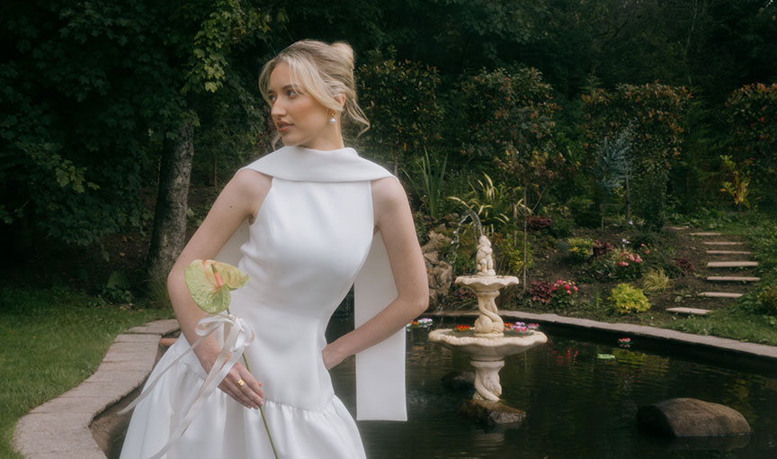 Bride in a modern white gown holding a single flower, standing beside a garden pond and stone fountain.