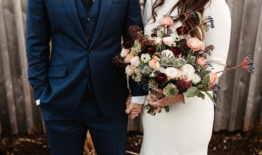 a person in a dark blue suit standing next to a person in an ivory dress holding a bouquet of flowers