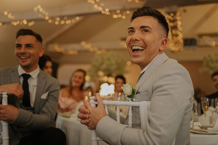 Two People Wearing Grey Suits Seated At A Table Smile with twinkling lights and people out of focus in background