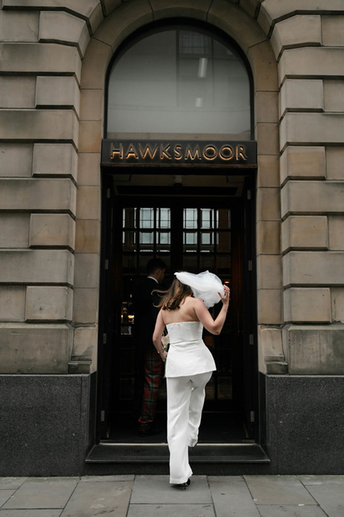 Couple entering Hawksmoor Edinburgh on their wedding day, with one wearing a white trouser suit and veil.
