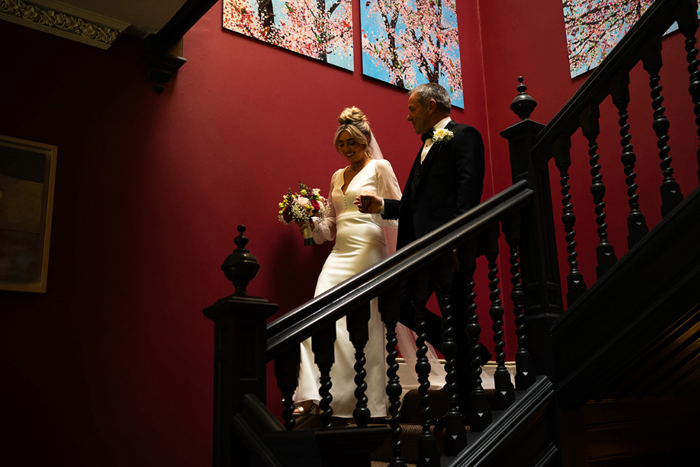 Bride and father walk downstairs against red walls