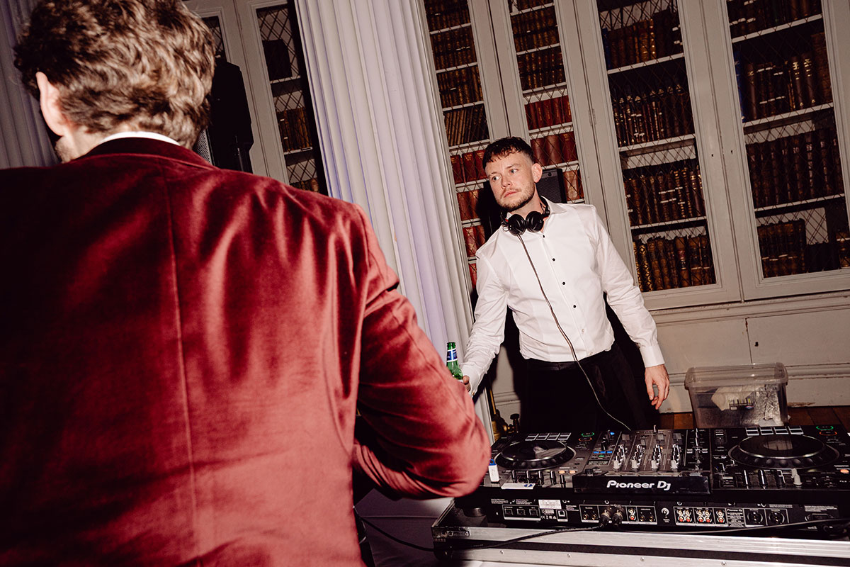 Groom standing behind a DJ booth at the Signet Library, wearing headphones and holding a drink as guests gather on the dancefloor